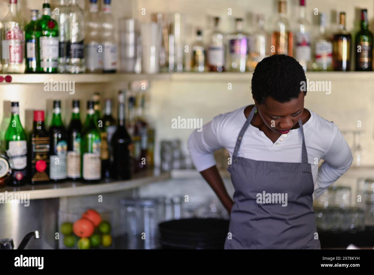 Il barista concentrato che indossa un grembiule si trova di fronte a una varietà di bottiglie in un bar pieno Foto Stock