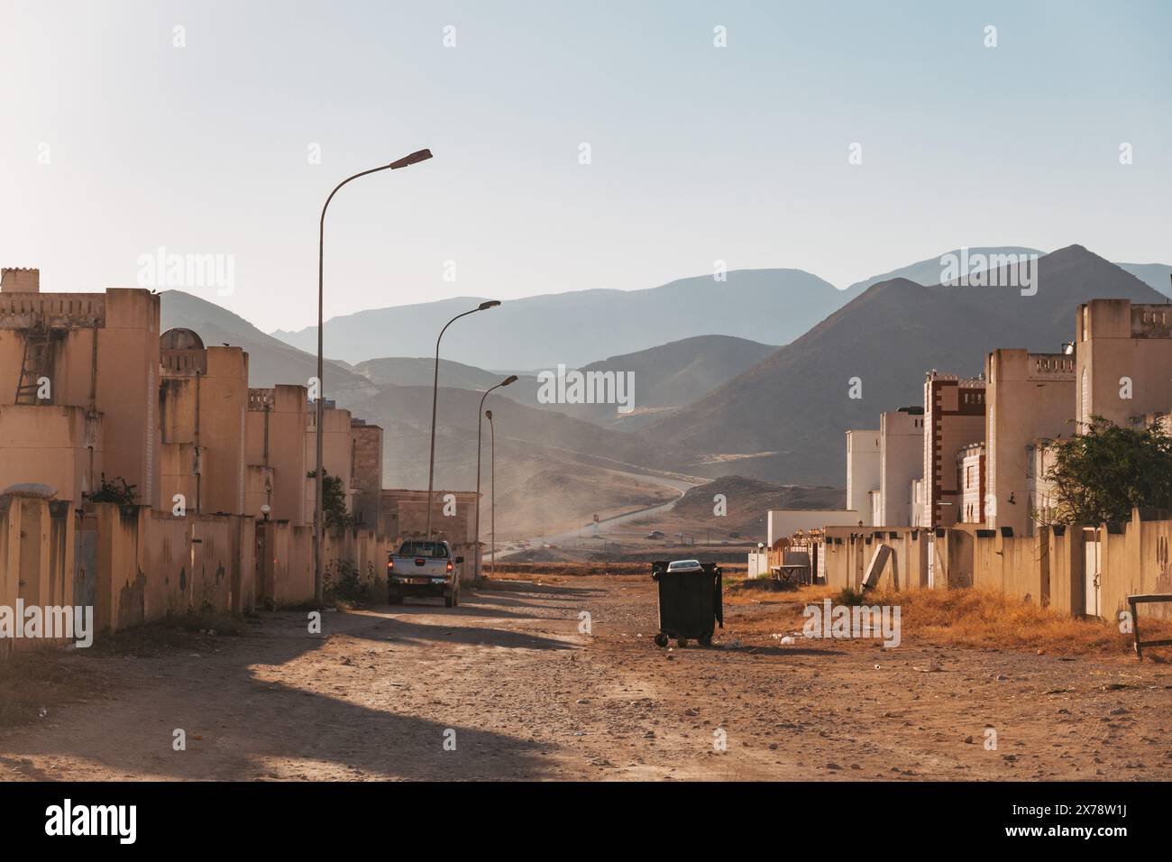 Strada residenziale a Mughsail, Oman, con montagne sullo sfondo e una strada polverosa fiancheggiata da case e lampioni. Foto Stock