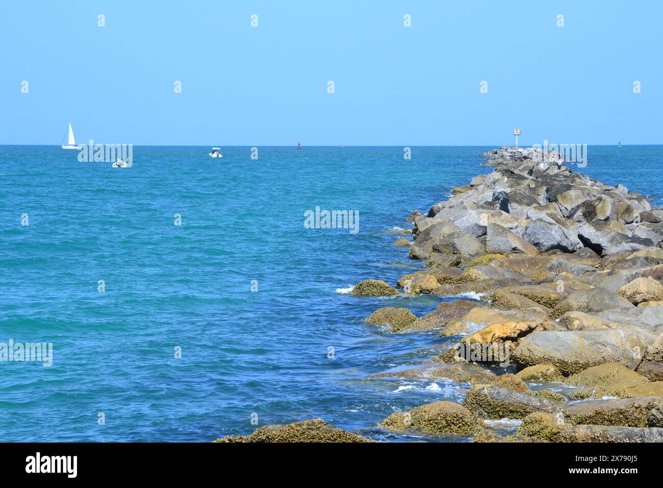 Un molo roccioso si estende nel mare turchese, con varie barche che punteggiano l'orizzonte a Ponce Inlet Beach, Florida. Foto Stock