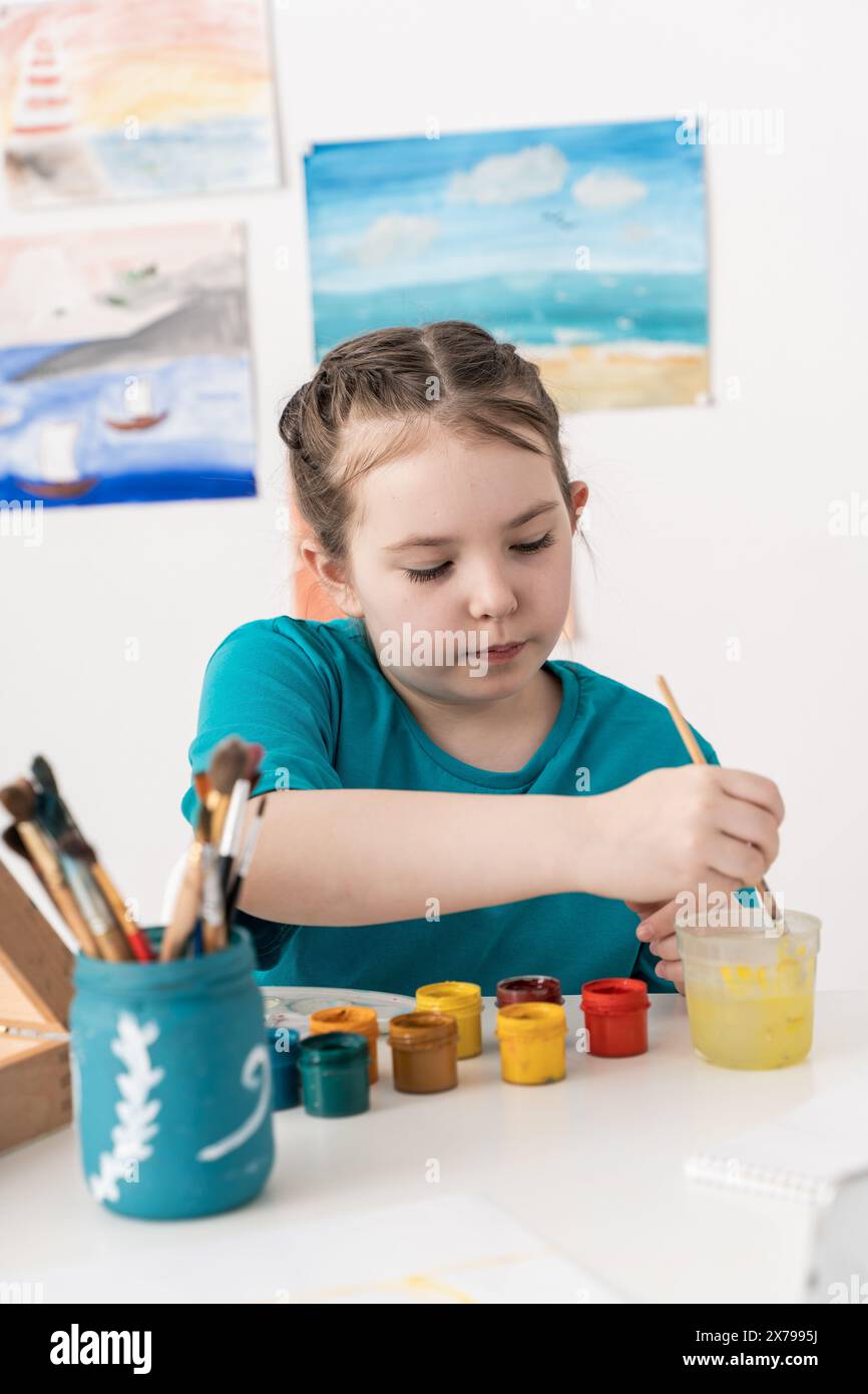 Una bambina disegna. Una giovane artista immerge un pennello nell'acqua mentre si siede sullo sfondo di un muro con i suoi disegni. Messa a fuoco selezionata. Alto Foto Stock