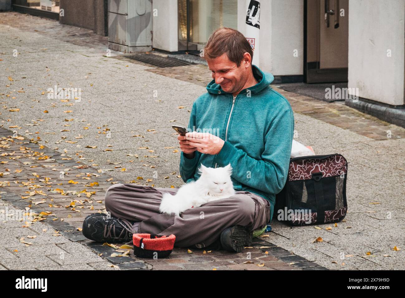 Un uomo sorridente siede a gambe incrociate su una strada di Lussemburgo con un gatto bianco in grembo, guardando il suo telefono, accanto a un portabagagli e a una ciotola per donazioni. Foto Stock