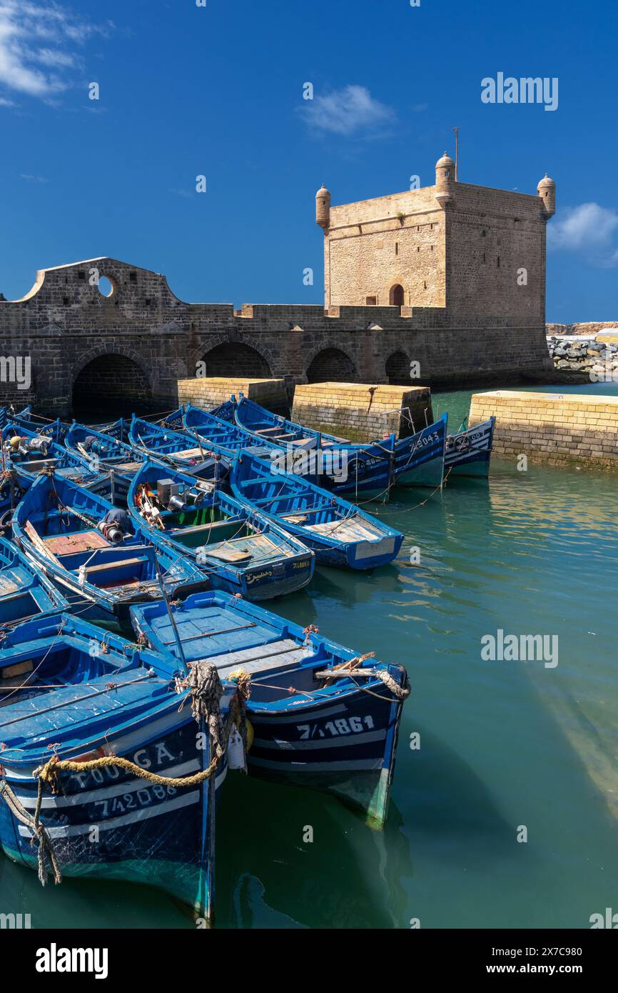 Essaouira, Marocco - 26 marzo 2024: La torre di guardia Borj el Barmil e le colorate barche da pesca blu nel vecchio porto di Essaouira Foto Stock