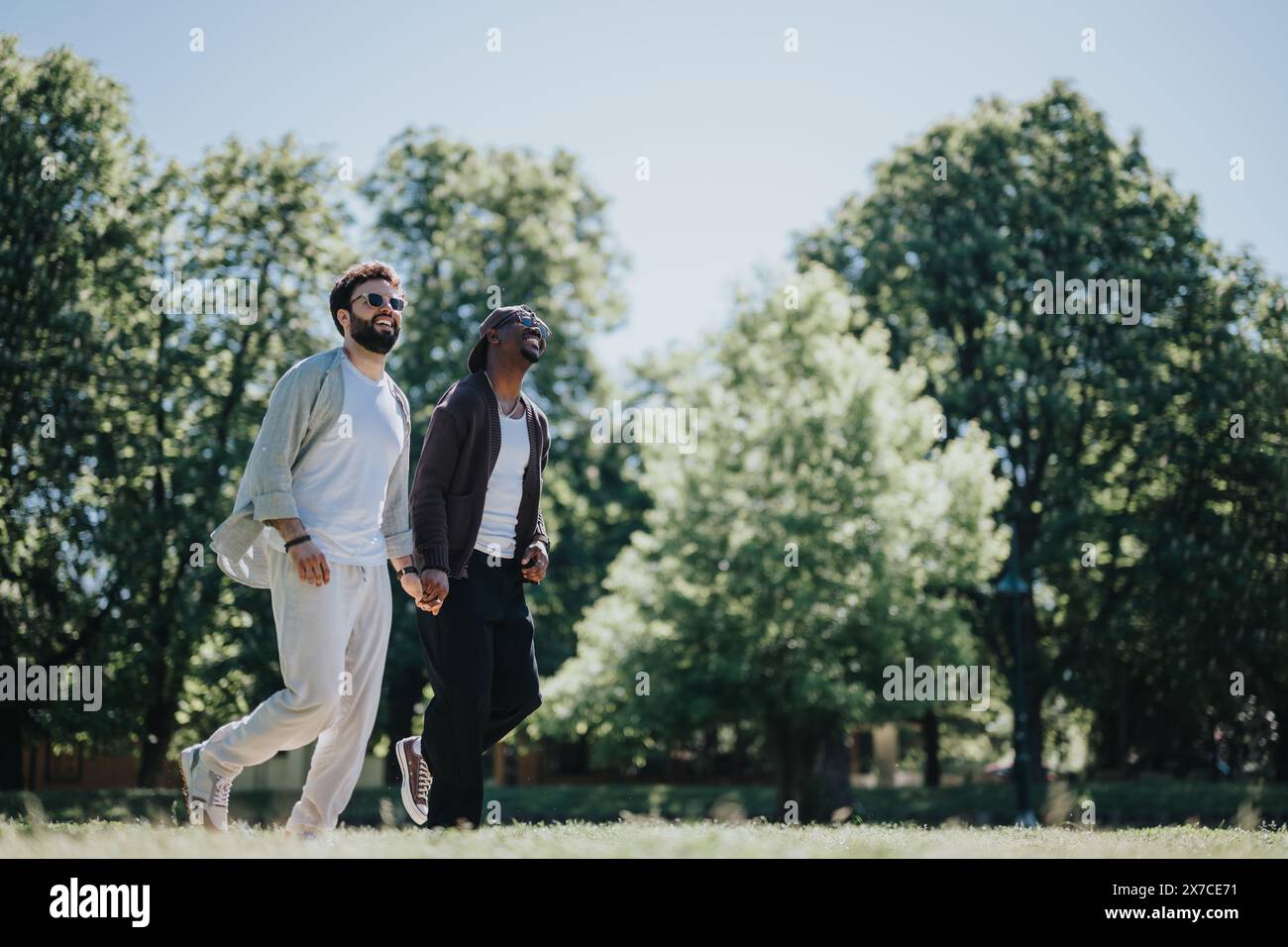 Coppia gay multirazziale che si gode una giornata di sole insieme nel parco, tenendosi per mano e sorridendo Foto Stock