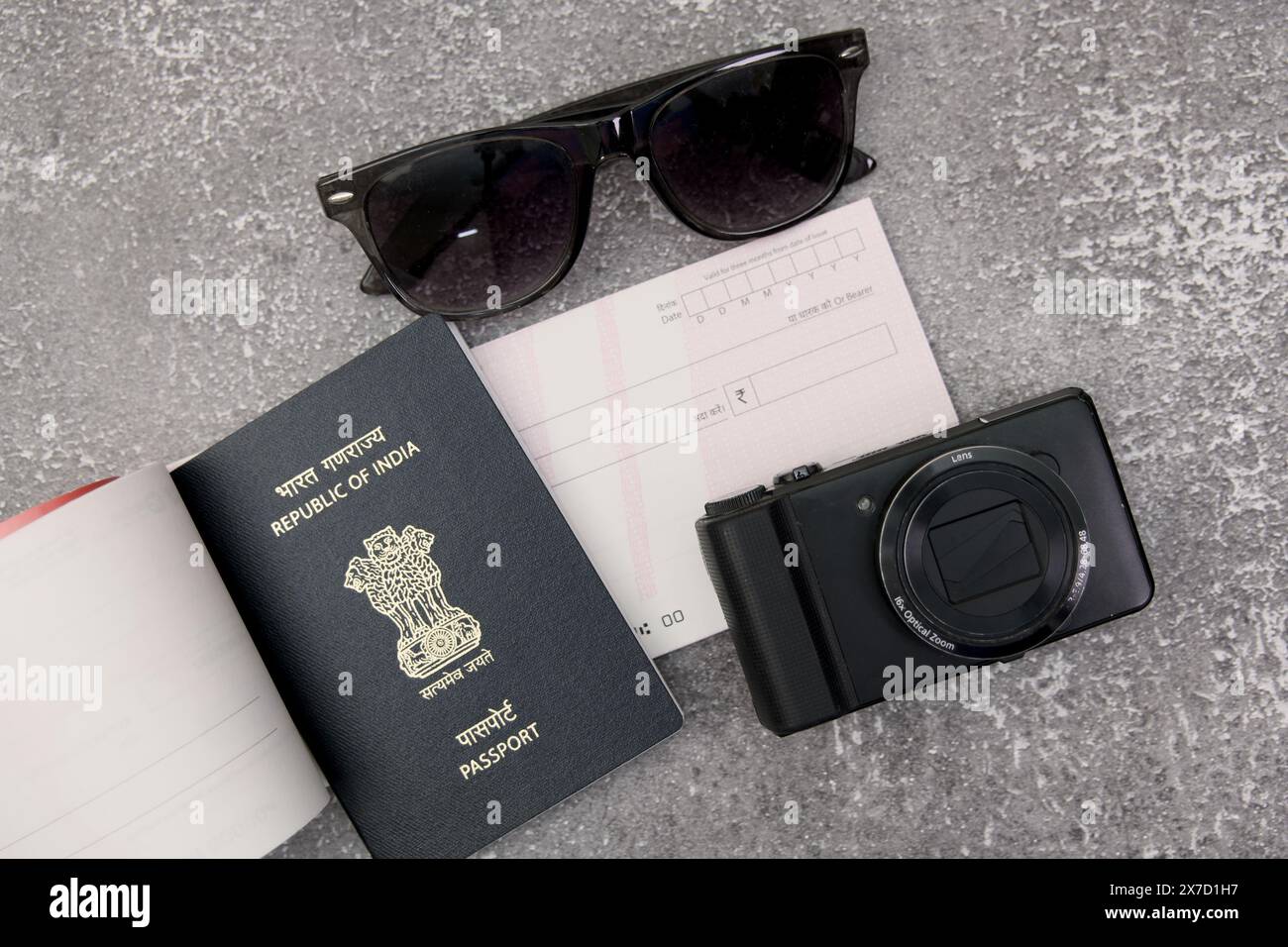 Questa immagine mostra i must di viaggio: Passaporto indiano, macchina fotografica, occhiali da sole e un assegno, tutti disposti su una superficie piatta in pietra grigia. Foto Stock