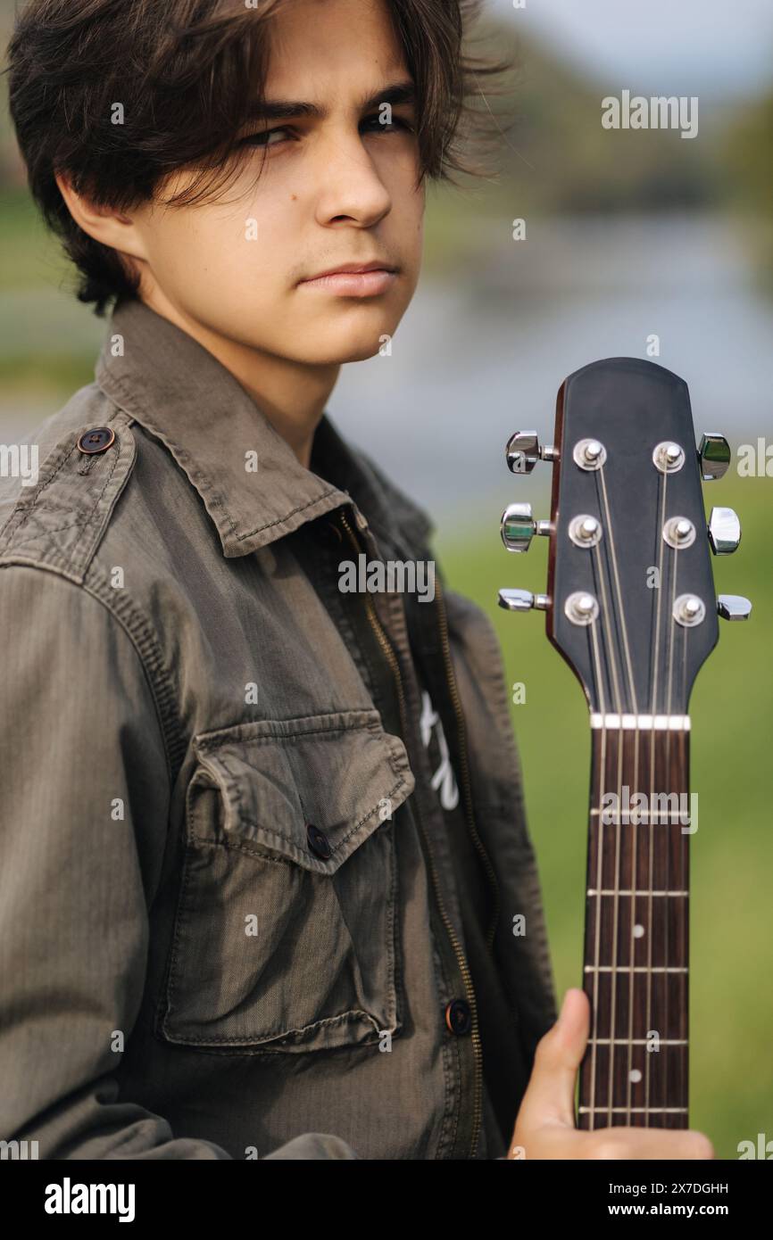 Ritratto di moda di capelli lunghi ragazzo tenere chitarra acustica. Ragazzo giovane che si prepara a suonare la chitarra all'aperto Foto Stock
