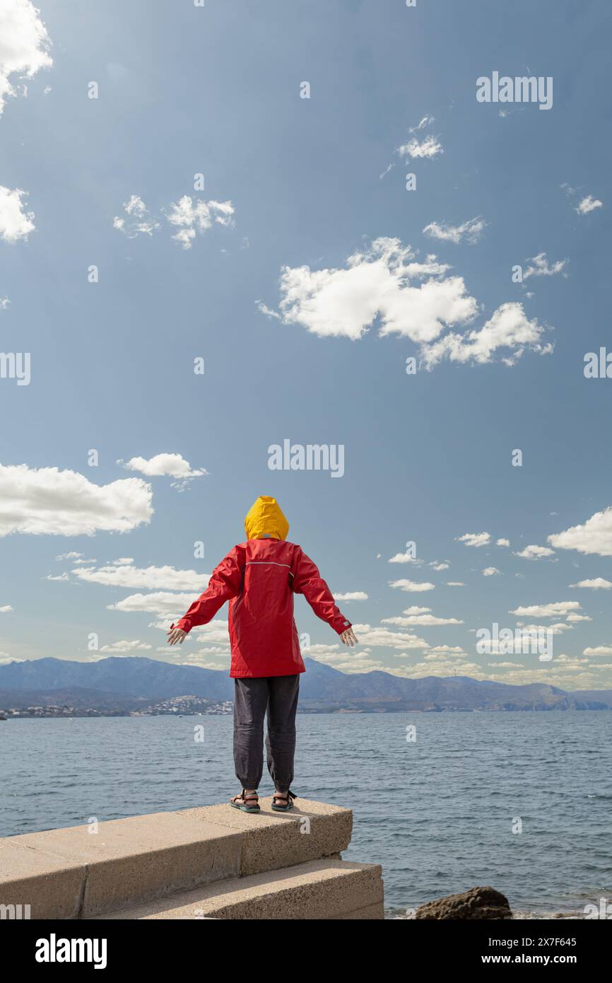 persona di fronte al mare con una tempesta sopra la testa con grandi nuvole grigie, giacca rossa con cappuccio giallo, braccia sollevate in segno di protesta Foto Stock