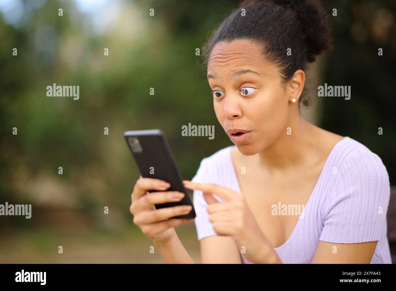 Una donna nera stupita che controlla il cellulare fuori in un parco Foto Stock