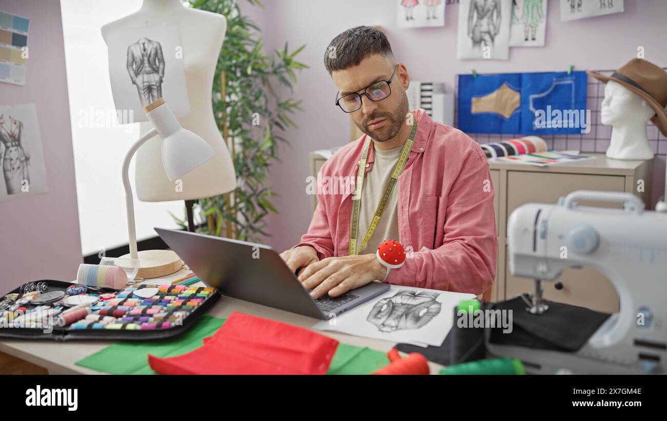 Uomo ispanico con occhiali e barba che lavora su un portatile in un colorato negozio di sartoria circondato da tessuti e attrezzature da cucito. Foto Stock