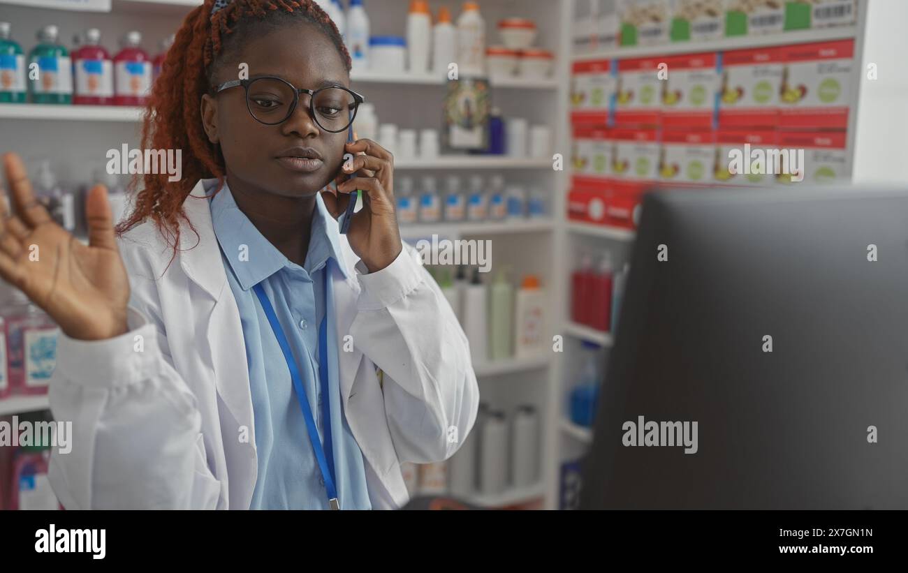Una giovane farmacista afroamericana che lavora in multitasking in una farmacia mentre è al telefono con scaffali di prodotti sullo sfondo. Foto Stock