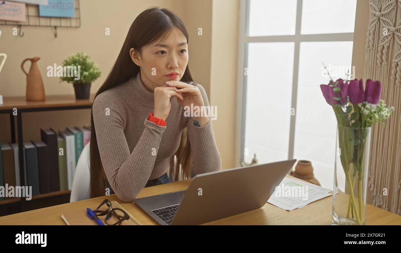 Una contemplativa donna cinese siede in casa in un ufficio domestico con un notebook, un paio di occhiali e fiori viola. Foto Stock