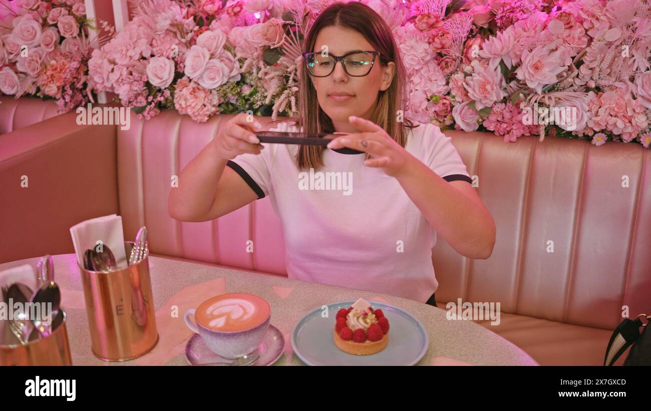 Donna adulta che fotografa i dessert in un caffè rosa dal design floreale Foto Stock