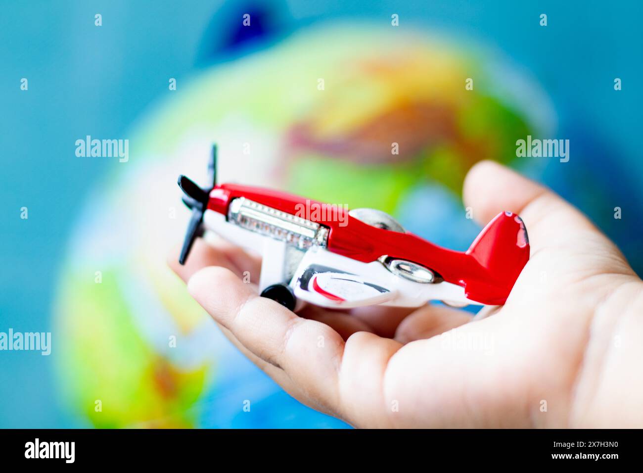 Mano del bambino che tiene un aereo giocattolo con un'elica nel palmo. Aereo giocattolo rosso davanti allo sfondo del modello mondiale. Concetto di idea aeronautica. Tour mondiale. Foto Stock