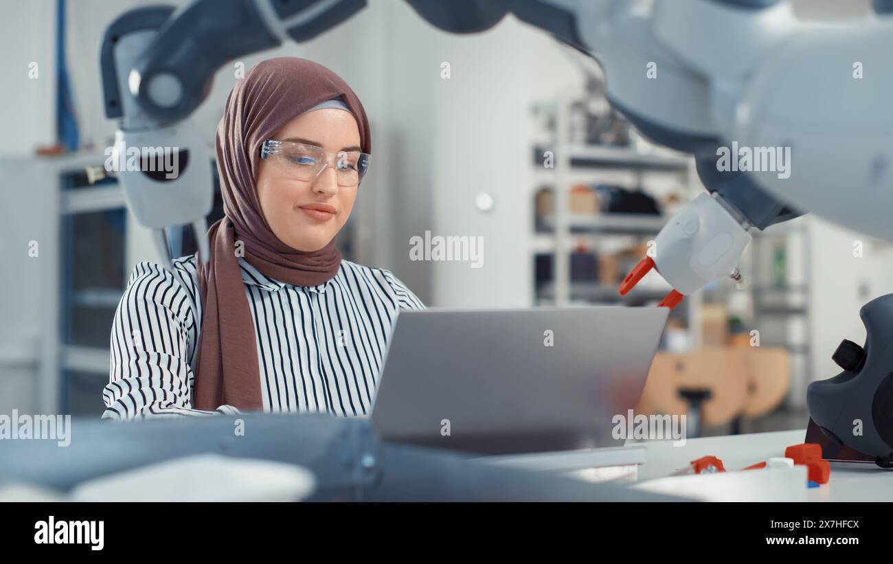 Ingegnere del Medio Oriente in Hijab conduce un'analisi approfondita del braccio robot e dei programmi su laptop in Modern Office. Ottimizzazione dello stabilimento di produzione autonomo per i prodotti High Tech. Foto Stock