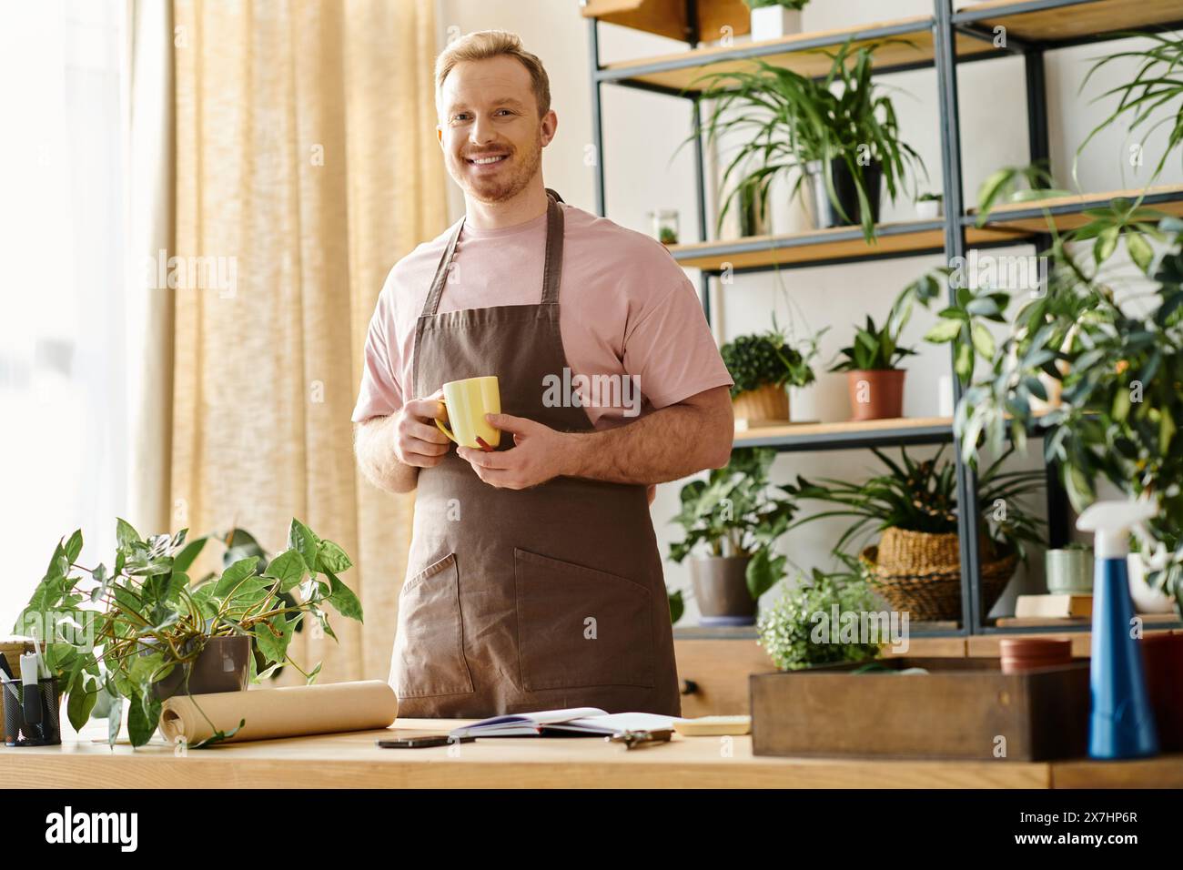 Un bell'uomo in grembiule si gode una tazza di caffè in un negozio di piante, incarnando l'essenza di possedere una piccola impresa. Foto Stock