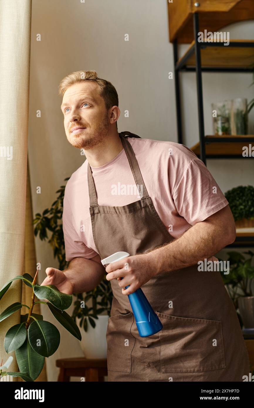 Un bell'uomo in un grembiule che tiene una bottiglia spray blu in un negozio di piante, riflettendo il fascino di possedere una piccola attività floreale. Foto Stock