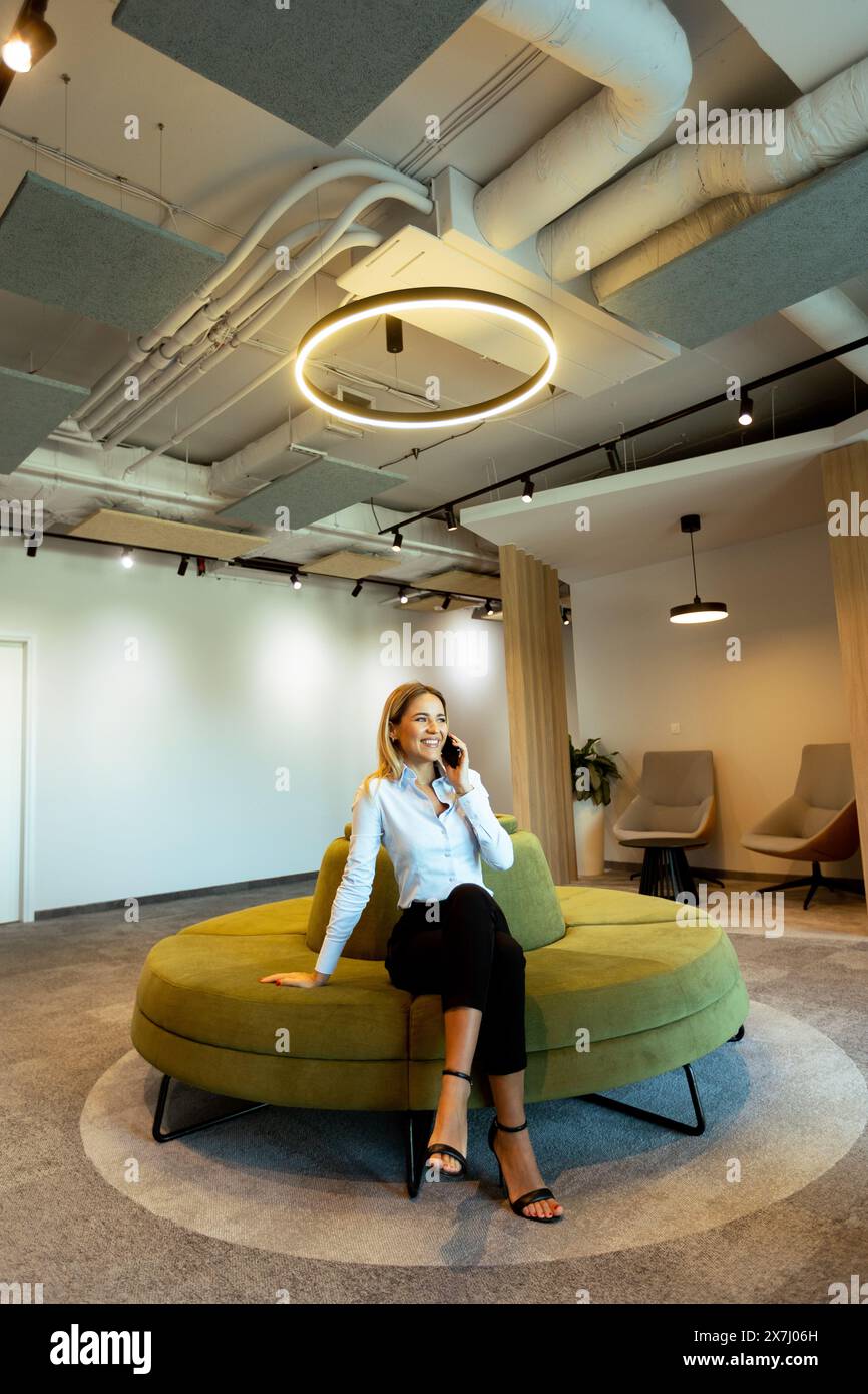 Donna professionale in un informale abbigliamento d'affari, seduto su un divano verde contemporaneo, impegnata in una telefonata all'interno di uno spazio ufficio elegante e moderno Foto Stock