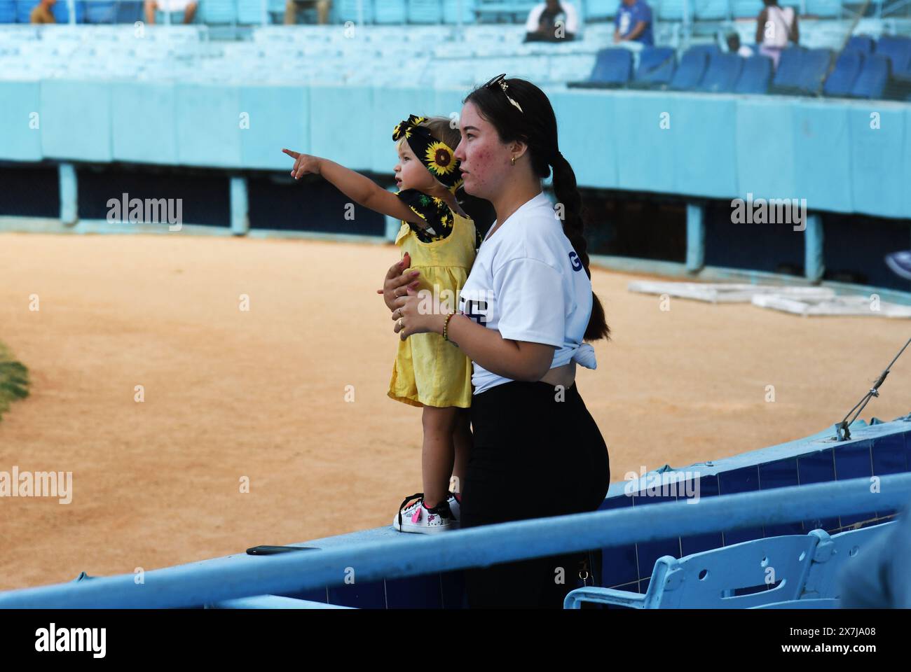 Una ragazza cubana entusiasta di vedere un'immagine di suo padre sul jumbotron a una partita di baseball a l'Avana, Cuba, e indica che la madre la tiene e guarda. Foto Stock