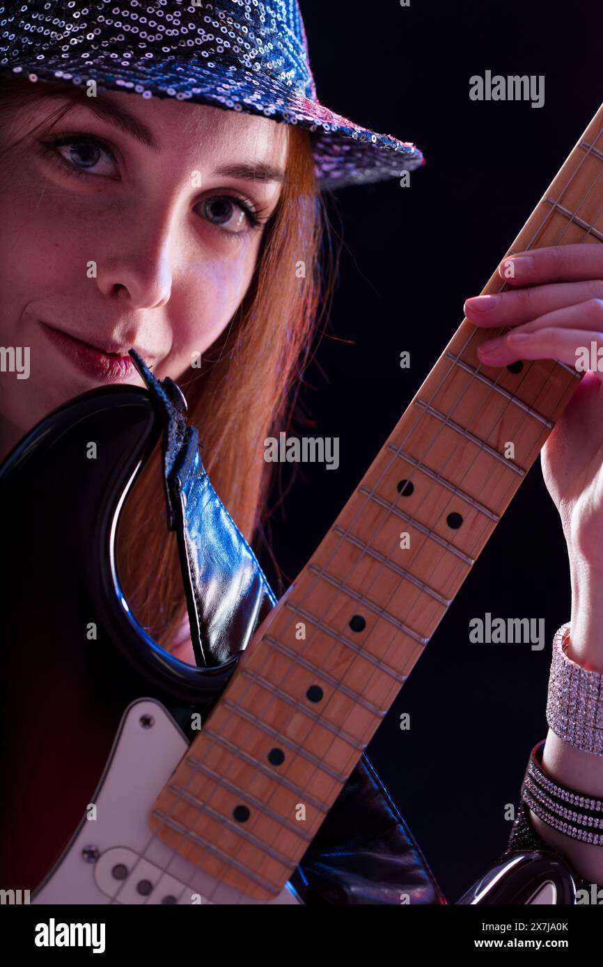 giovane donna con i capelli di auburn, vestita con un cappello scintillante e una maglia scura, suona una chitarra elettrica con un sorriso. La sua felice performance mette in risalto i suoi pas Foto Stock