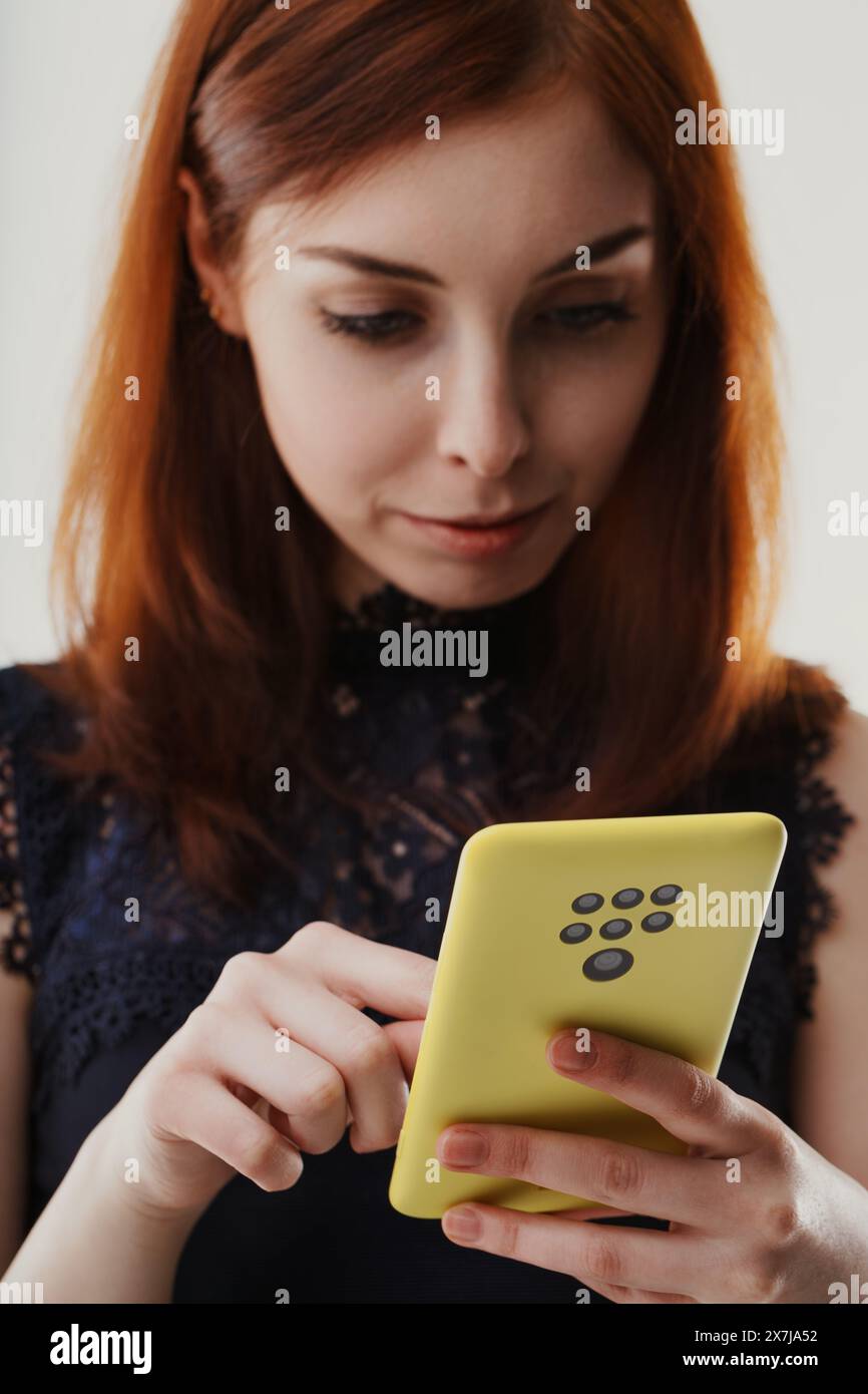 La giovane donna con i capelli di auburn guarda con attenzione il suo smartphone giallo, che ha un numero insolitamente elevato di telecamere sul retro. Indossa un pizzo scuro Foto Stock