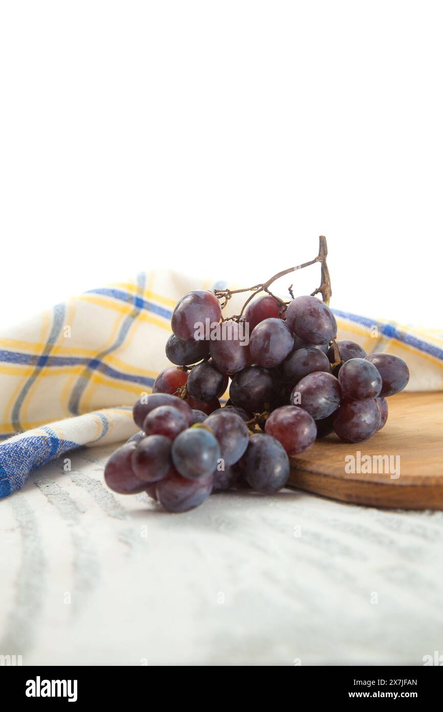 Tagliere con grande mazzo di uva blu scuro maturo su bianco. Uva da vino viola con asciugamano da cucina blu su fondo di legno bianco. Foto Stock