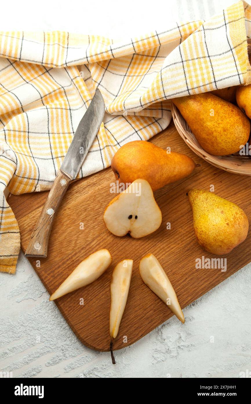 Tagliere in legno con tagliere interi, metà e fette di pere mature e succose, cesto di vimini con pere e asciugamano da cucina giallo su fondo in legno bianco Foto Stock