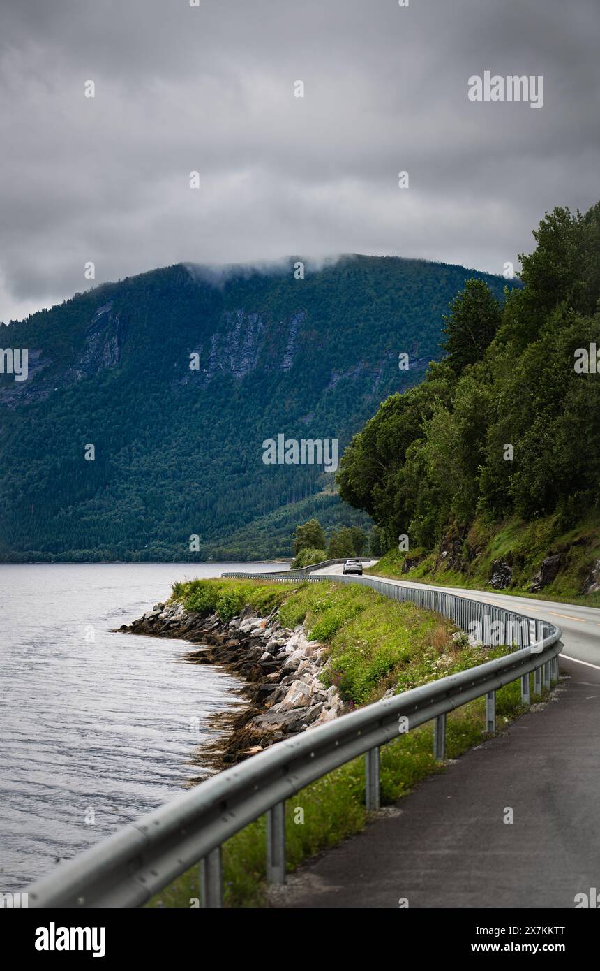 Un'auto solitaria percorre la Norwegian National Road Highway 70 accanto all'Alvundfjord, circondata da vegetazione lussureggiante, montagna in lontananza sotto il cielo nuvoloso Foto Stock