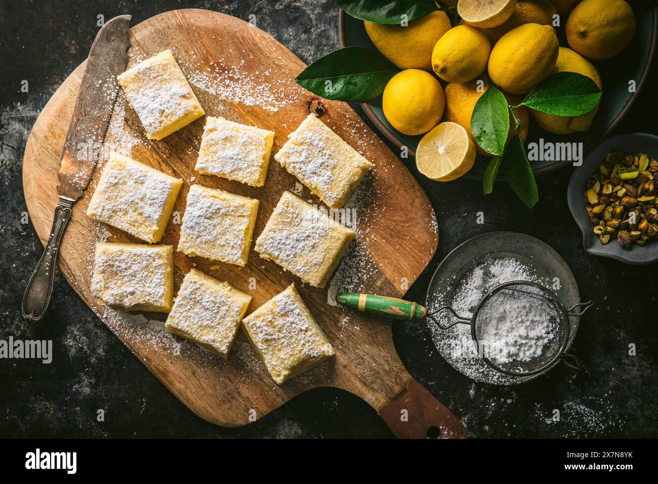 Torta al limone, tagliata a quadrati, su un tagliere di legno con coltello per torte, zucchero a velo e ciotola di limoni freschi Foto Stock