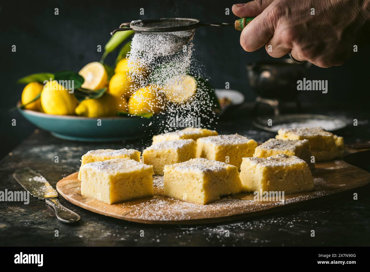 Colpo d'angolo laterale con la mano del maschio che spolverano zucchero a velo sui quadrati di torta al limone, ciotola di limone in BG Foto Stock