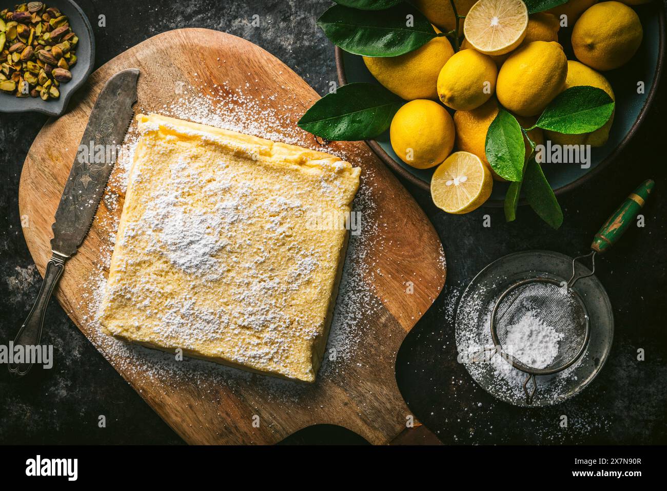 Torta al limone, non affettata, su un tagliere di legno con coltello per torte, zucchero a velo e ciotola di limoni freschi Foto Stock