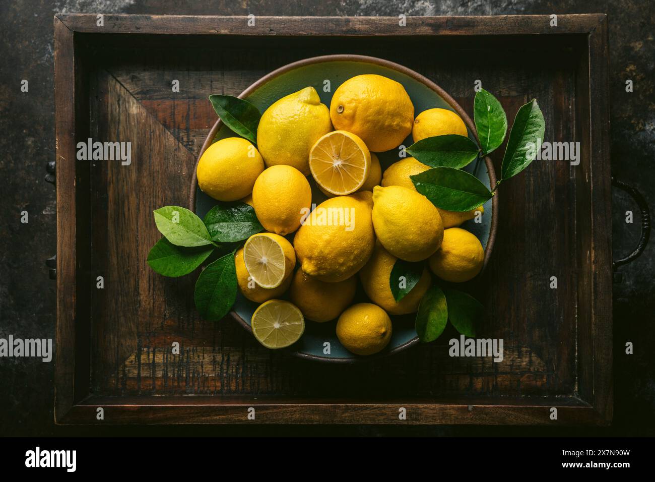 Varietà di limoni di Lisbona e Meyer, alcuni tagliati, con foglie fresche, preparati in una ciotola di ceramica blu. Incastonato in una scatola di legno rustico Foto Stock