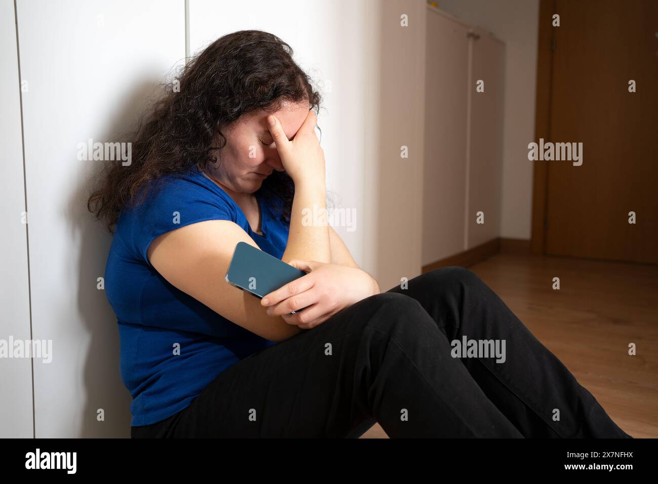 Una donna triste che regge un telefono cellulare lamentandosi seduto sul pavimento di casa con una luce scura sullo sfondo Foto Stock