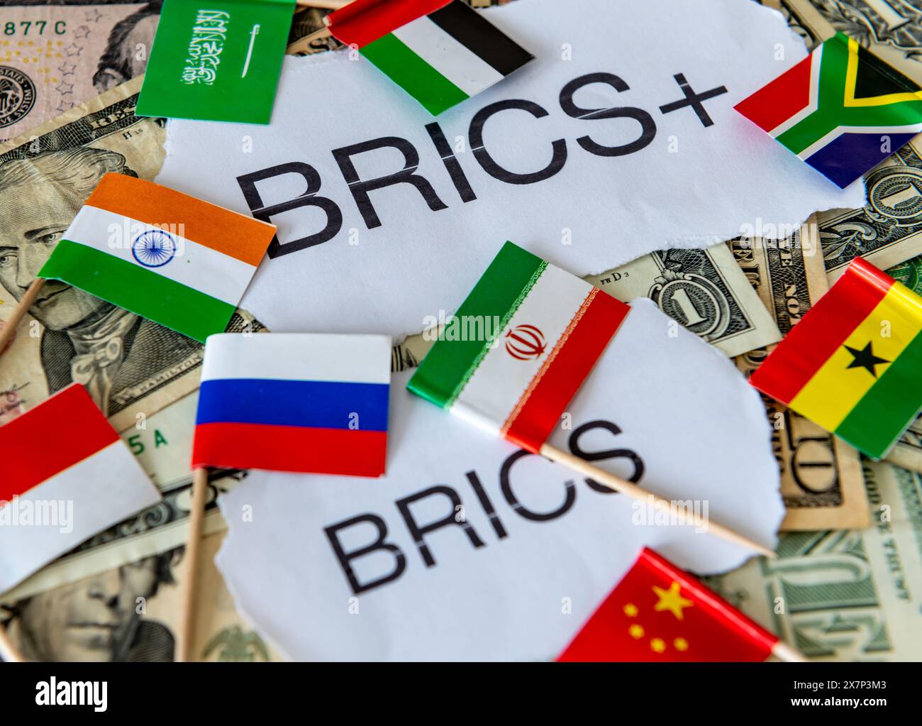 Le parole e le bandiere di alcuni dei paesi BRICS e BRICS+, oltre a una pila di banconote in dollari USA. Un concetto di dedollarizzazione. Foto Stock