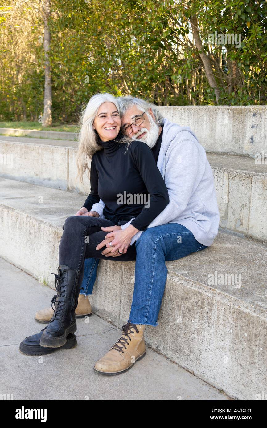 Una coppia anziana che mostra affetto mentre si siede insieme su gradini di cemento all'aperto, sorridendo e godendo la compagnia reciproca in un parco naturale Foto Stock