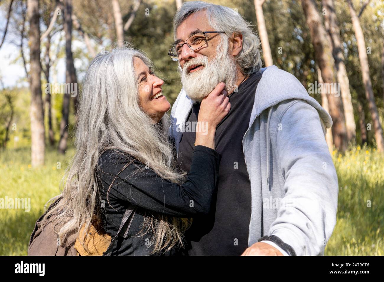 Un uomo e una donna senior si abbracciano gioiosamente in un parco illuminato dal sole, mostrando affetto e felicità nei loro anni d'oro, i loro sorrisi genuini e la loro atmosfera informale Foto Stock