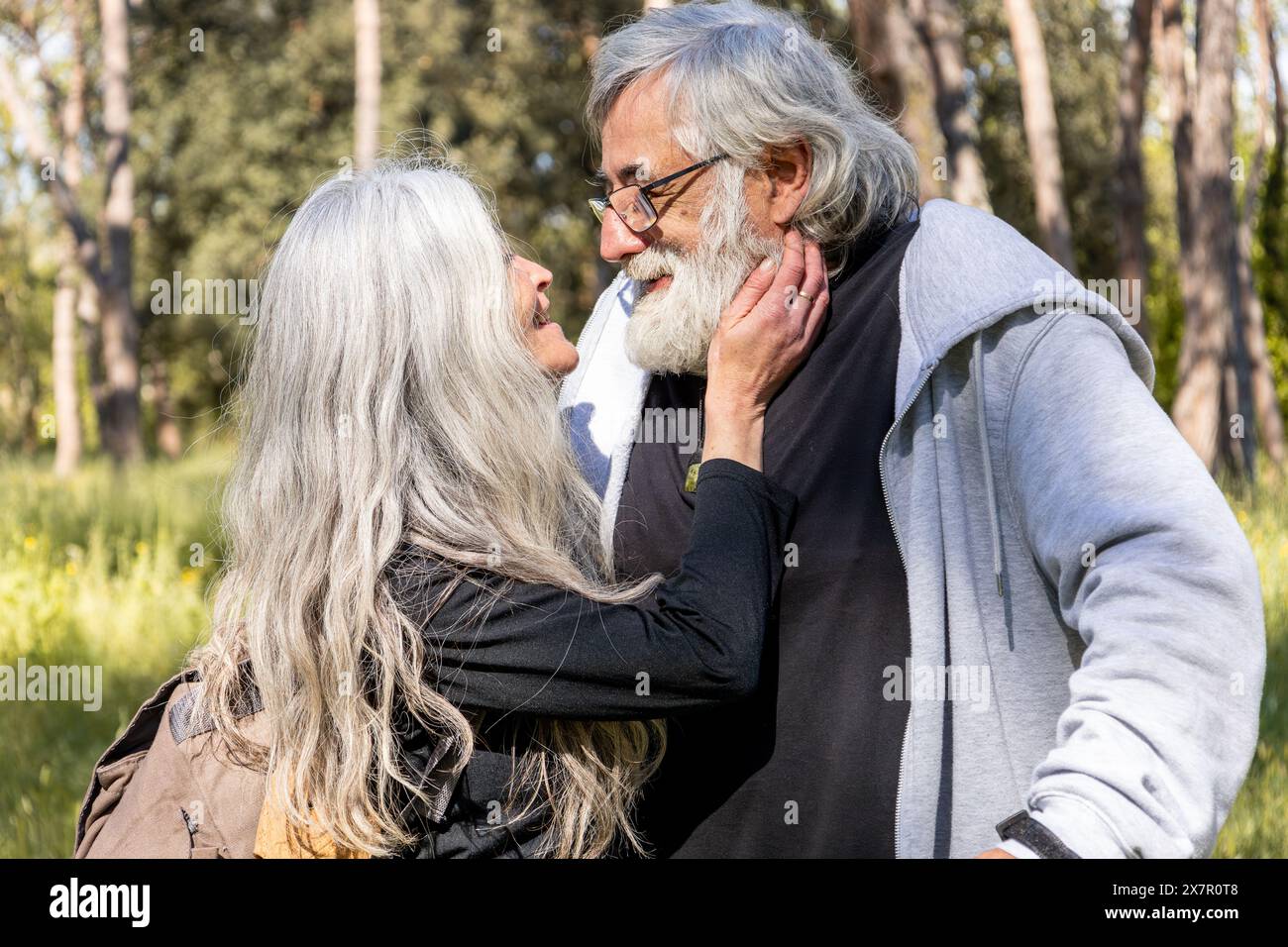 Un'affettuosa coppia senior che si guarda in un parco illuminato dal sole, con l'uomo che culla dolcemente il volto della donna, circondato da una vegetazione lussureggiante Foto Stock