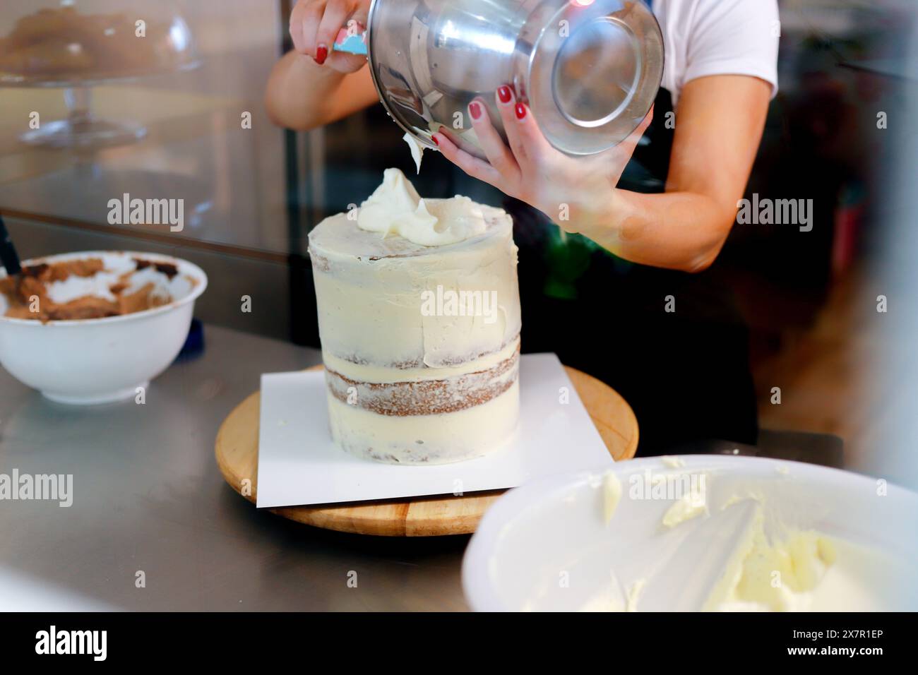 Un individuo aggiunge meticolosamente uno strato di glassa a una torreggiante torta a più strati utilizzando una spatola metallica, con ciotole di ingredienti nelle vicinanze Foto Stock
