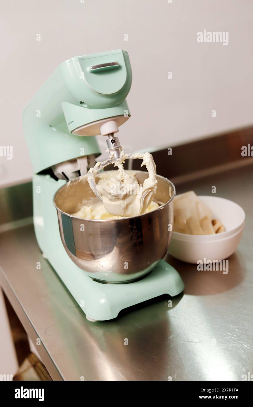 Un mixer a piedistallo verde menta monta la panna in un recipiente in acciaio inossidabile, essenziale per cuocere le torte, con ingredienti nelle vicinanze Foto Stock