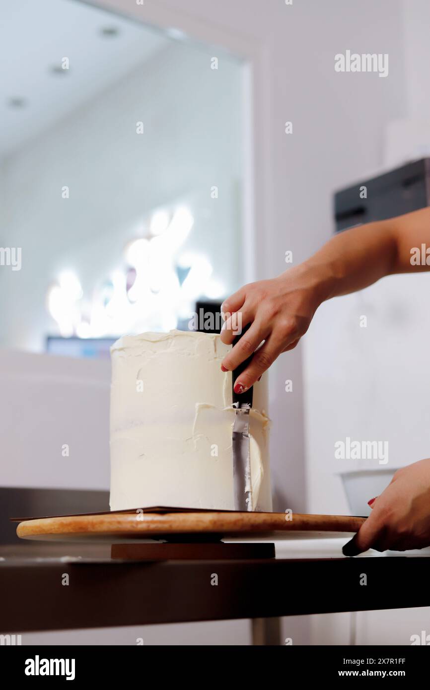 Una persona applica sapientemente una glassa liscia a una torta alta in una cucina moderna, dimostrando le tecniche di decorazione della torta Foto Stock