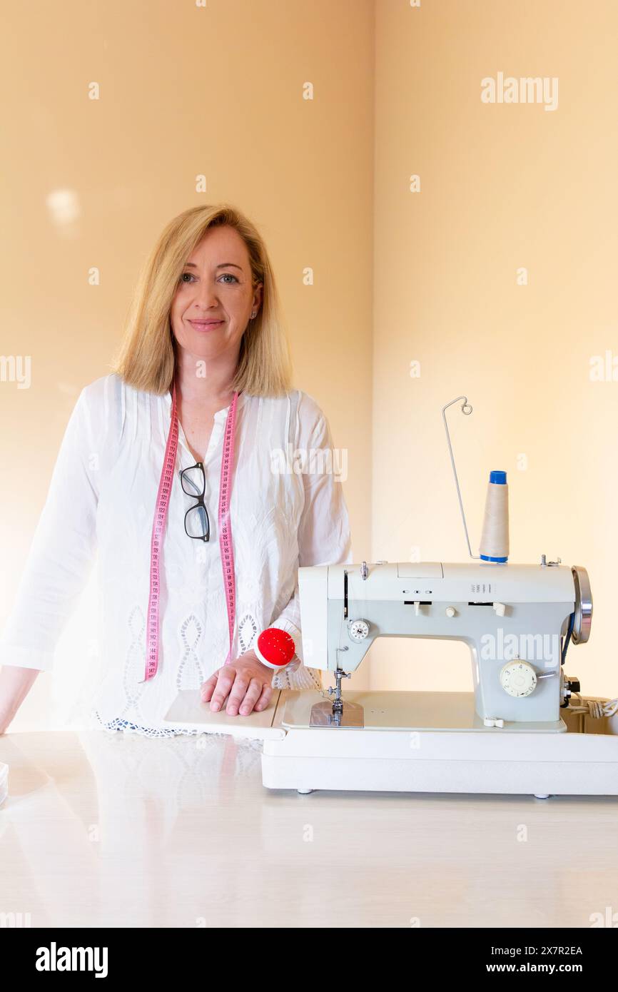 Una sarta professionista si trova orgogliosamente nella sua officina di cucito accanto a una robusta macchina da cucire con una bobina di filo blu e un cuscino rosso Foto Stock