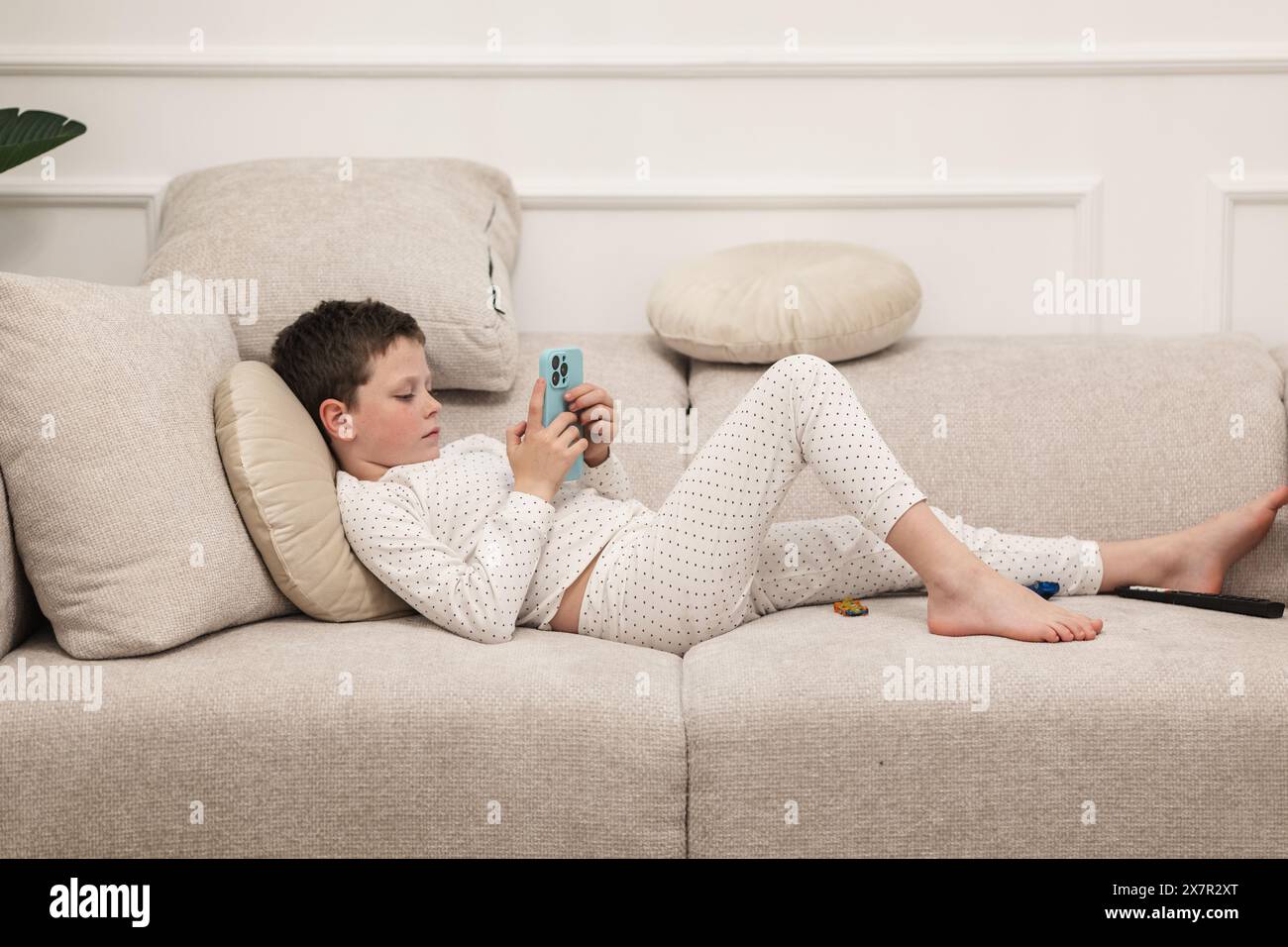 Un ragazzino sdraiato su un divano, concentrato sul suo smartphone, rivestito di pigiama bianco con puntini di polka neri. Foto Stock