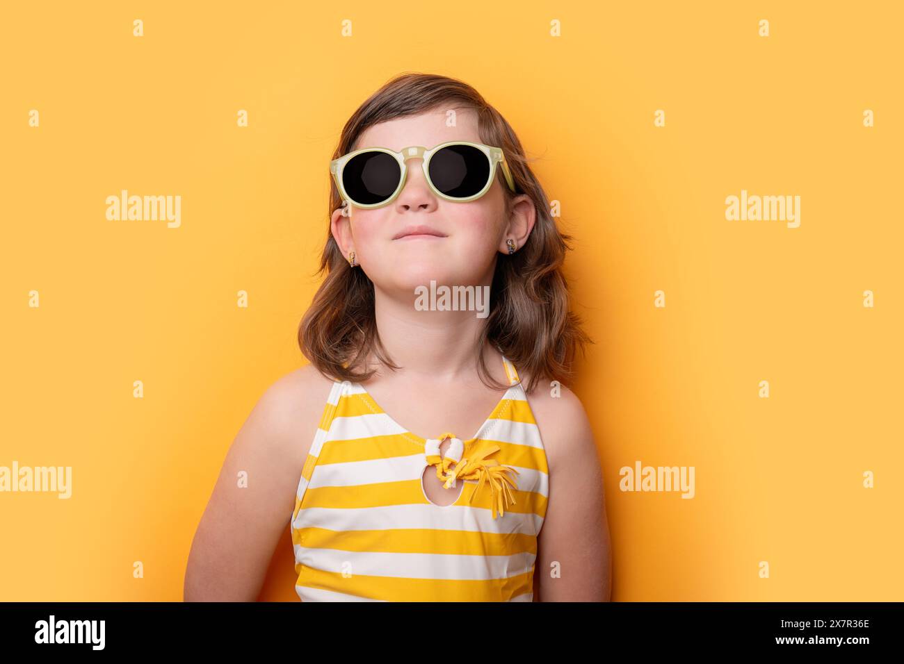 Una giovane ragazza sorride delicatamente indossando occhiali da sole oversize e un costume da bagno a righe giallo-bianche, davanti a un vivace sfondo giallo Foto Stock