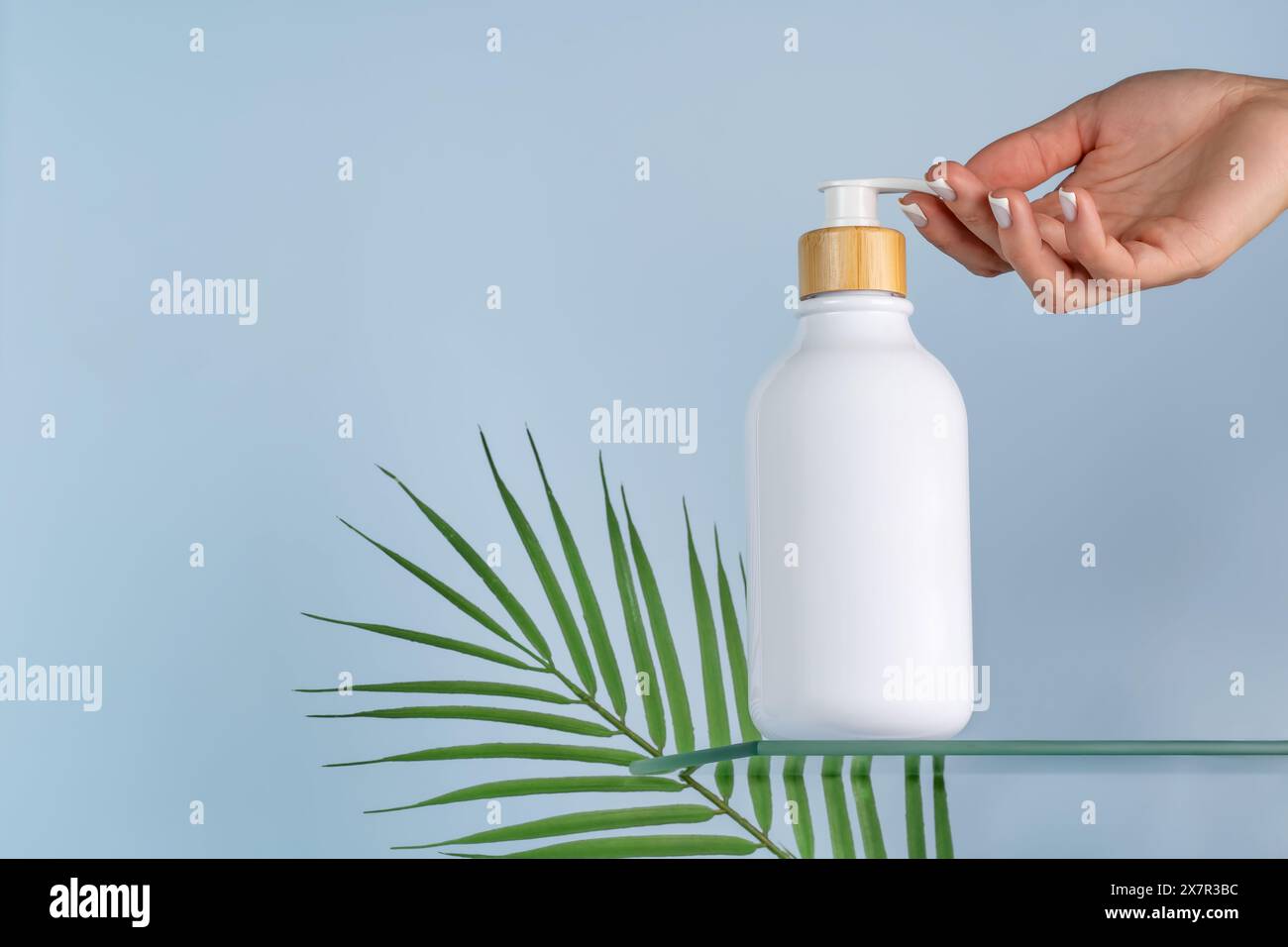 Una donna corta e irriconoscibile contiene un flacone bianco per cosmetici per la presentazione di un mockup su un sereno sfondo blu, con una fresca pubblicità a foglia verde Foto Stock