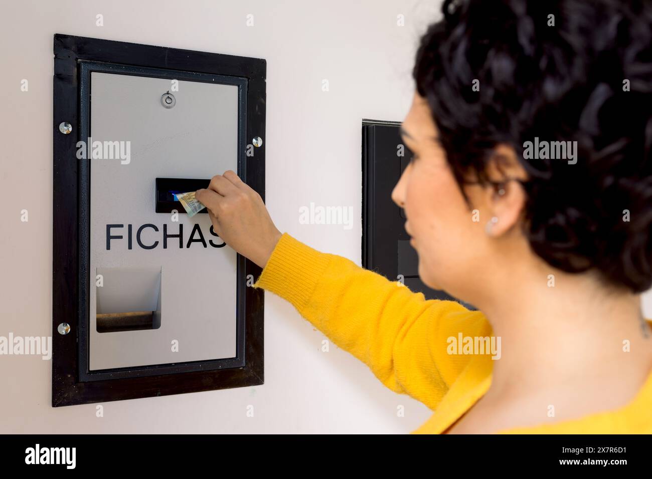 Una donna con un maglione giallo viene catturata mentre inserisce una banconota in un distributore automatico di gettoni, con la schiena rivolta verso la telecamera, in un ambiente interno Foto Stock