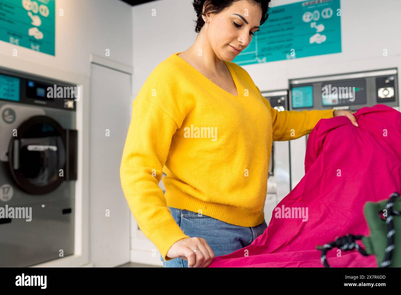 La vista laterale di una donna adulta concentrata consiste nello smistamento di un foglio rosa luminoso in una lavanderia pubblica con lavatrici sullo sfondo. Non guarda da t Foto Stock