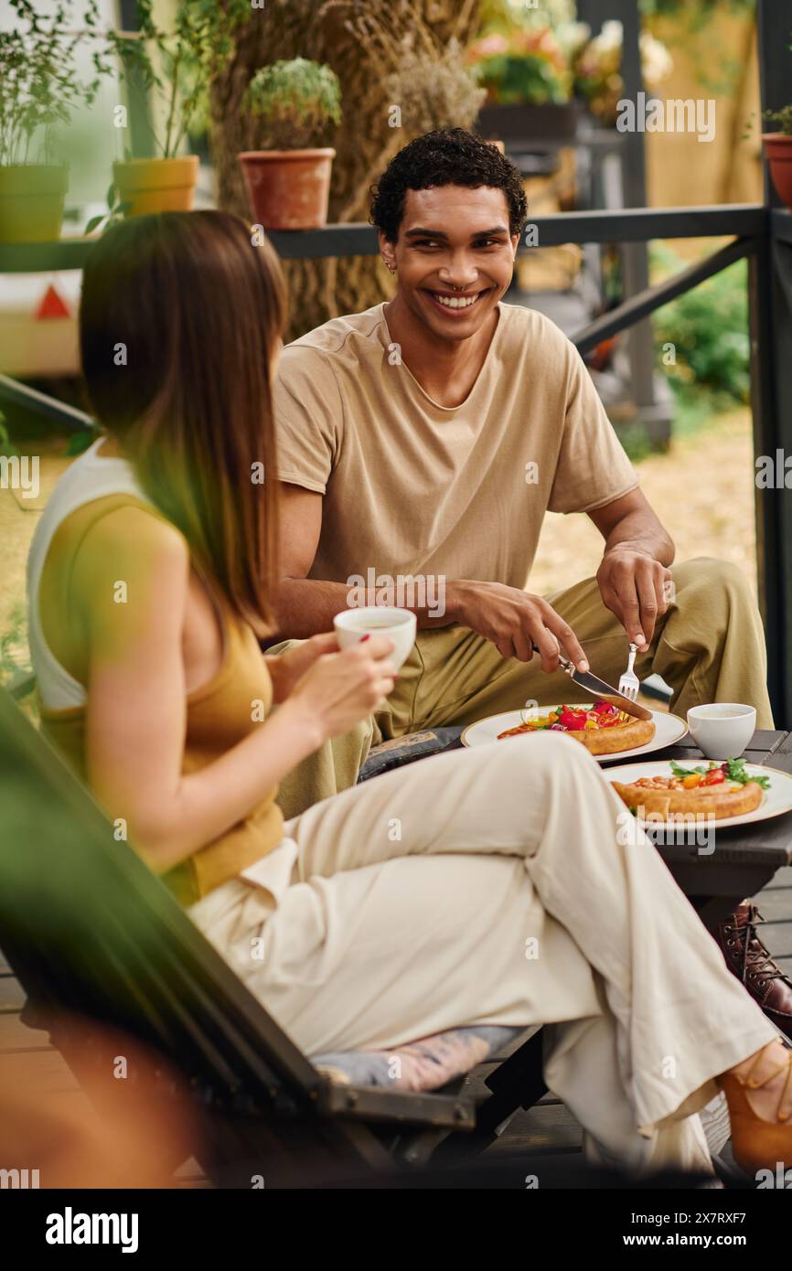 Una coppia interrazziale si diverte a fare un picnic su una panchina, condividendo un pasto insieme in un momento di romantico legame. Foto Stock