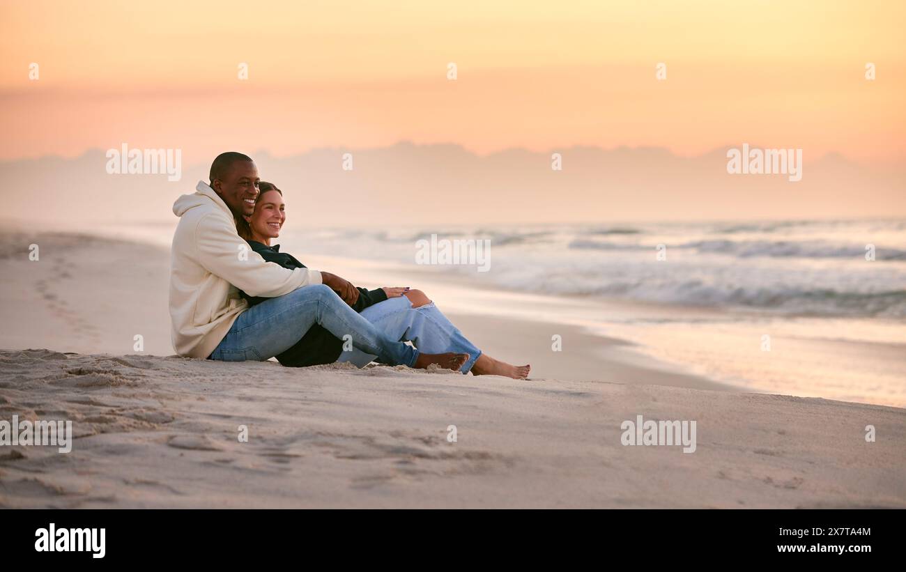 Una giovane coppia amorevole vestita in modo informale seduta sulla spiaggia che abbraccia all'alba Foto Stock