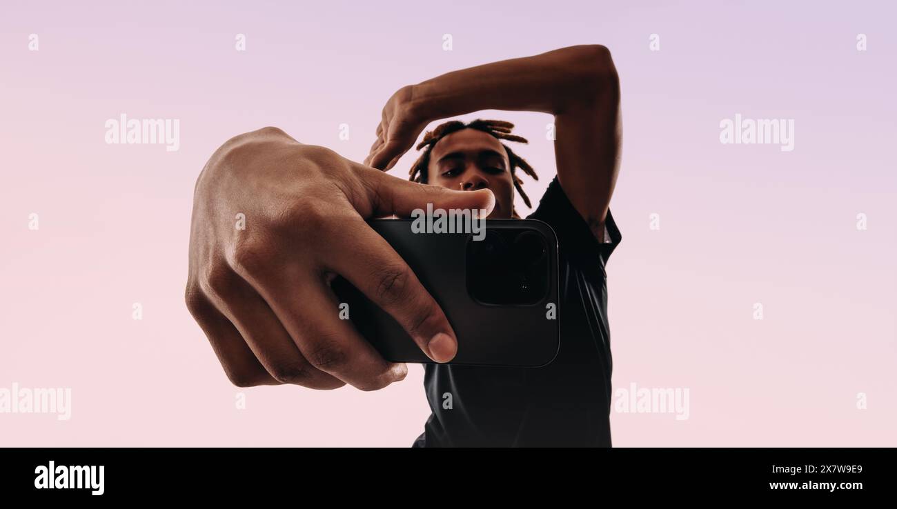 Un giovane maschio scatta con sicurezza un autoritratto con la fotocamera per selfie del suo smartphone. I suoi dreadlock gli inquadrano la faccia mentre tiene un dispositivo. Foto Stock