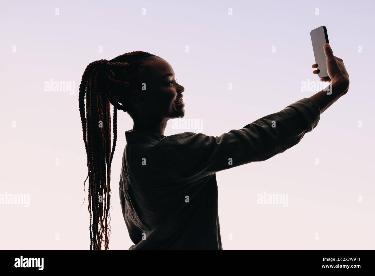Una donna sorridente sui vent'anni si trova in uno studio per un autoritratto. Ha un cellulare e si fa un selfie, mettendo in risalto la sua sicurezza e la sua vita Foto Stock