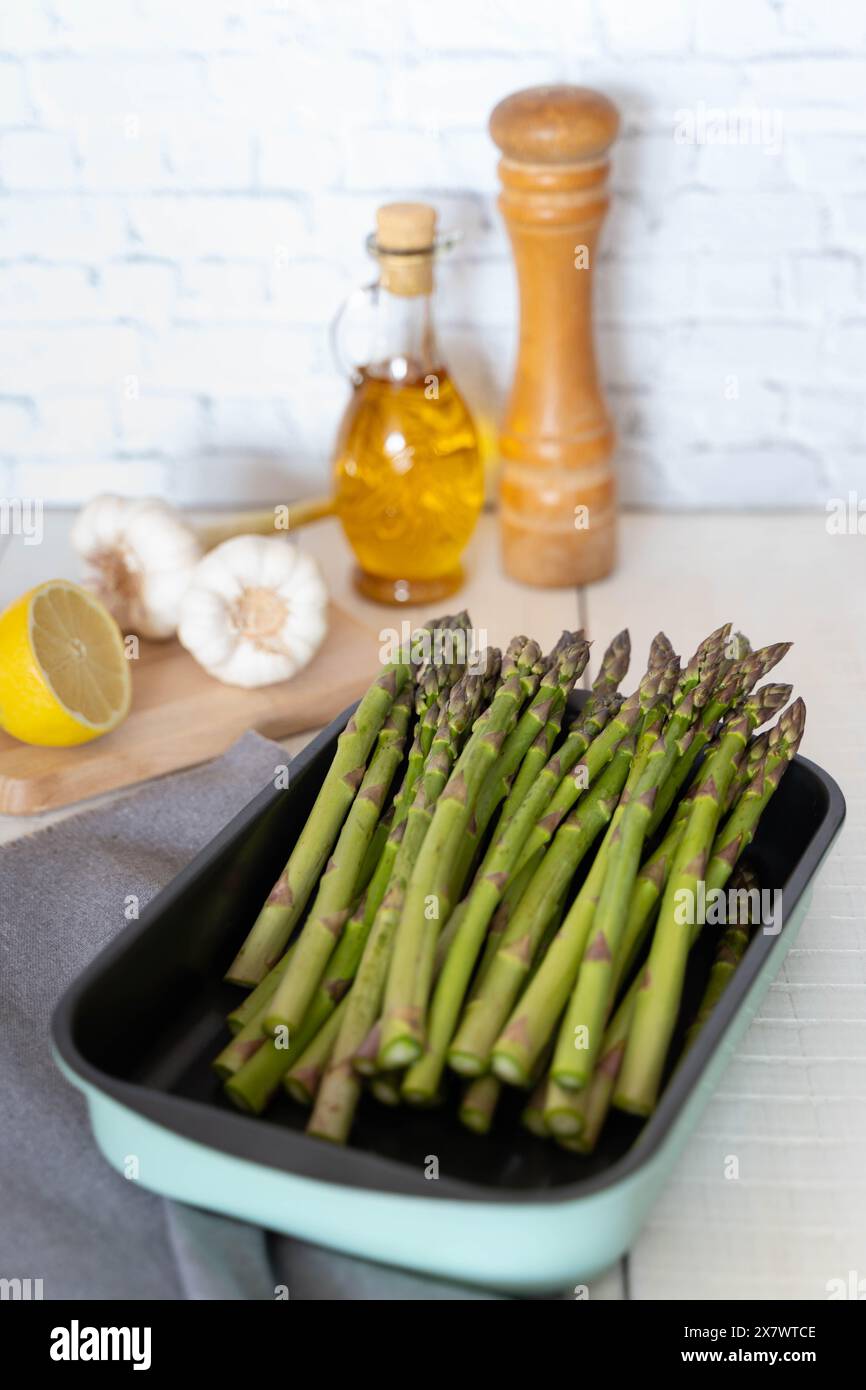 Asparagi verdi e croccanti disposti in vassoio da forno, preparati per la cottura. Mostrato contro un muro di mattoni, una bottiglia di olive e altri ingredienti Foto Stock