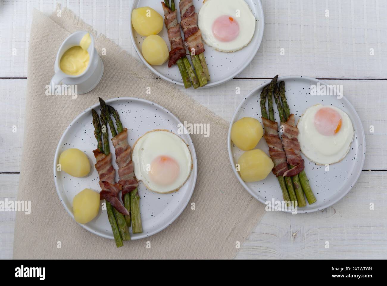 Asparagi verdi cotti al forno avvolti in pancetta e serviti con patate bollite e uova, su splendidi piatti bianchi su una superficie di legno chiaro Foto Stock